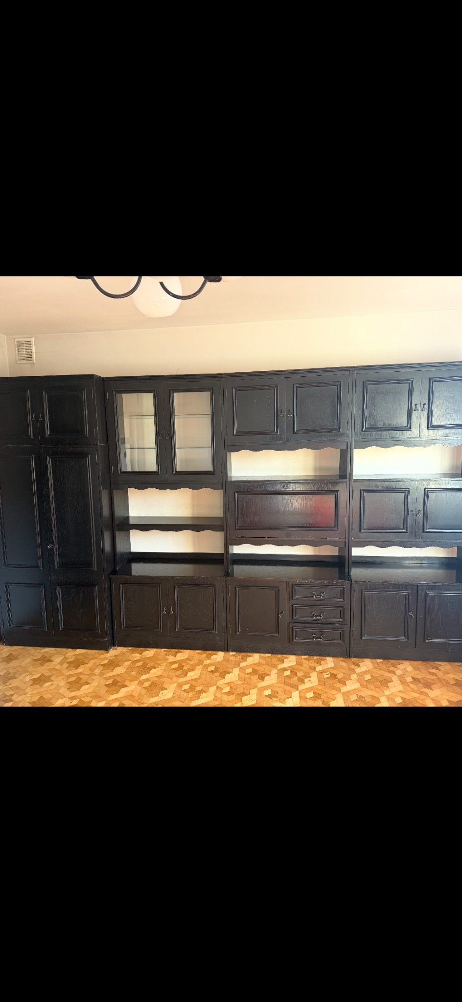 Ciemna, drewniana meblościanka z przeszklonymi witrynami i półkami, ustawiona na parkiecie. Widoczne detale wykończenia i oświetlenie sufitowe.