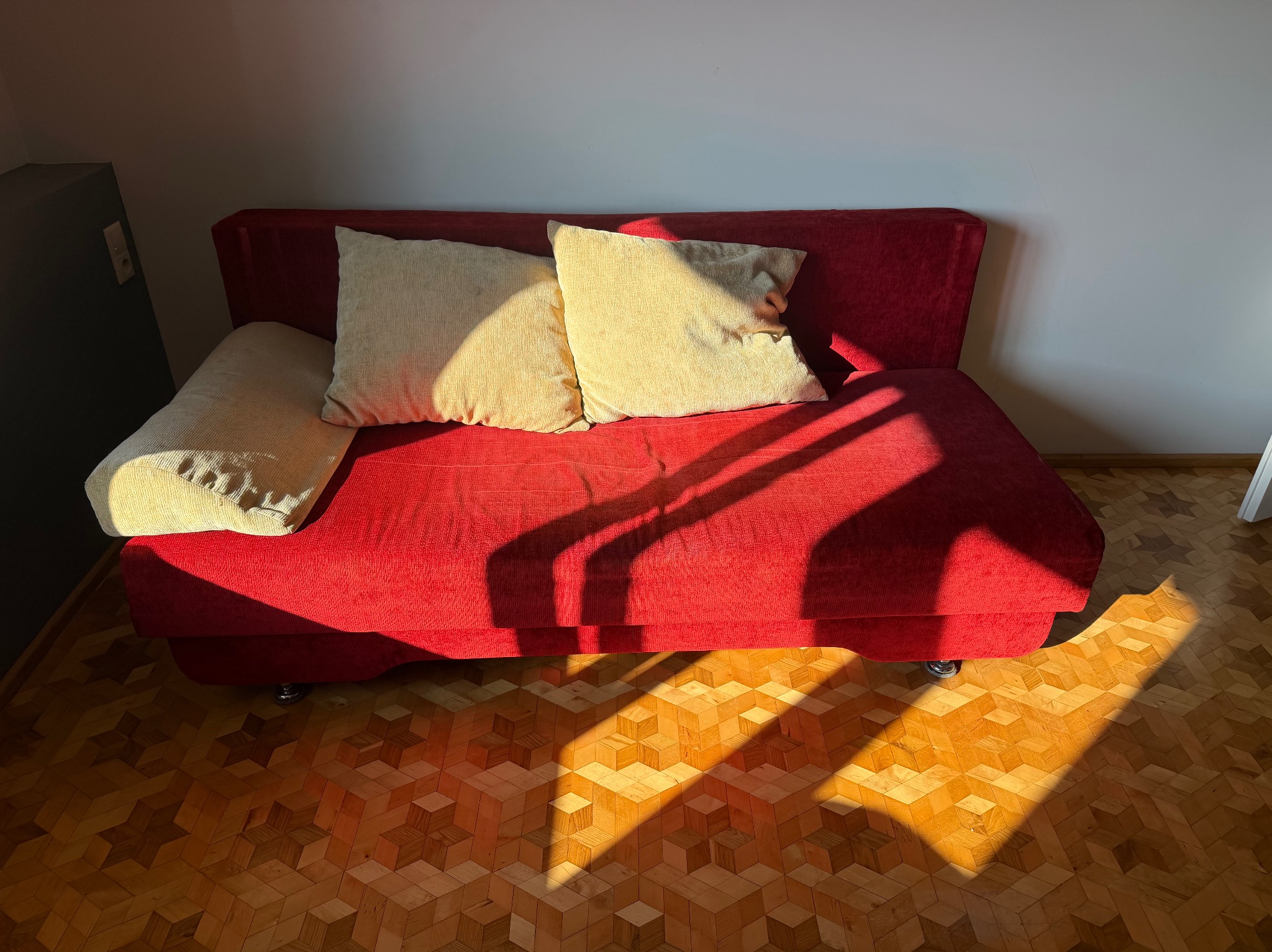 Czerwona sofa z beżowymi poduszkami w promieniach słońca na parkiecie w geometryczny wzór. Widok z góry, oświetlenie słoneczne tworzy mocne cienie.