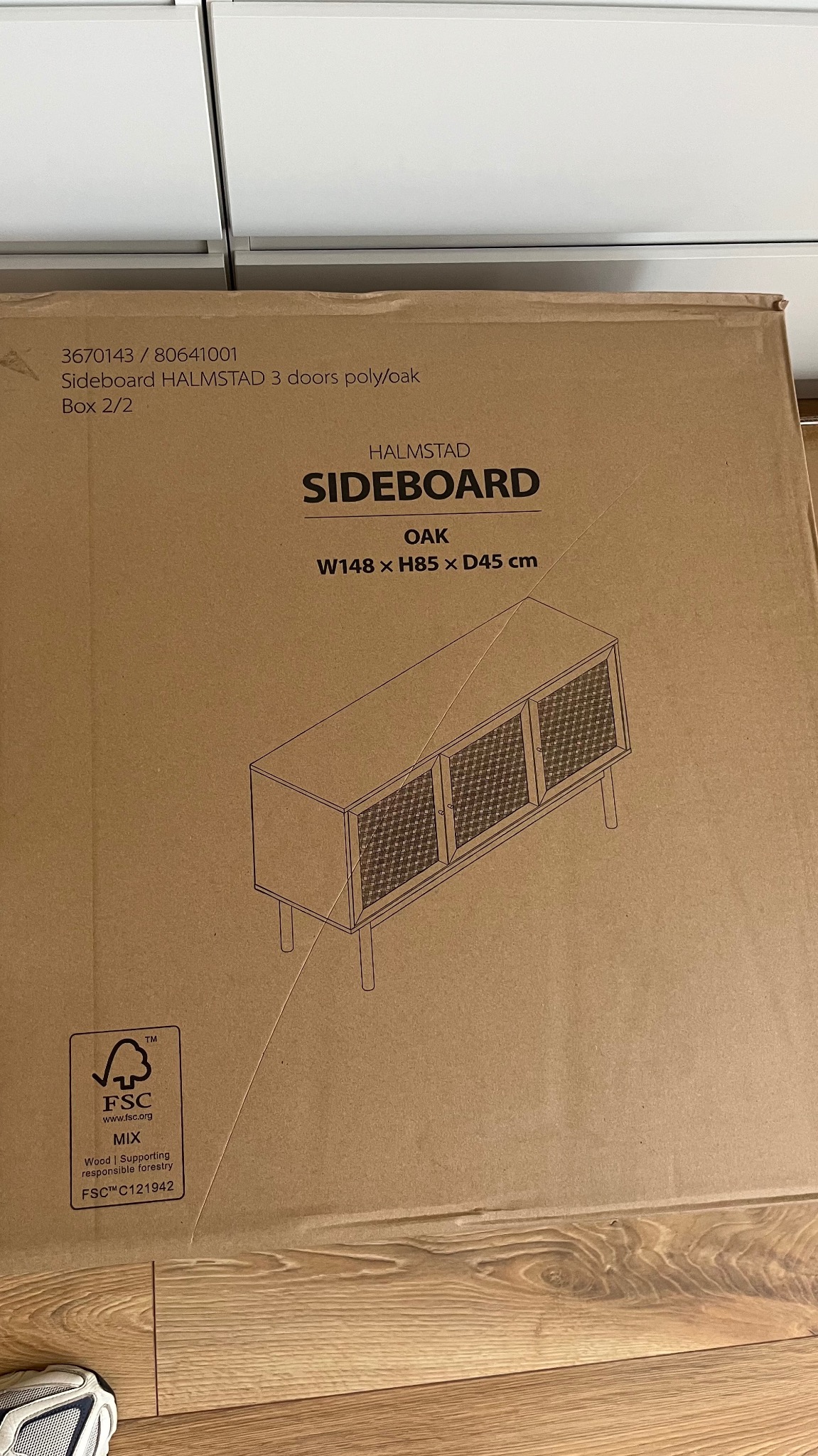 Kartonowe opakowanie mebla Halmstad sideboard z rysunkiem poglądowym, specyfikacją i logo FSC, leżące na podłodze. Widoczne fragmenty białych szafek w tle.