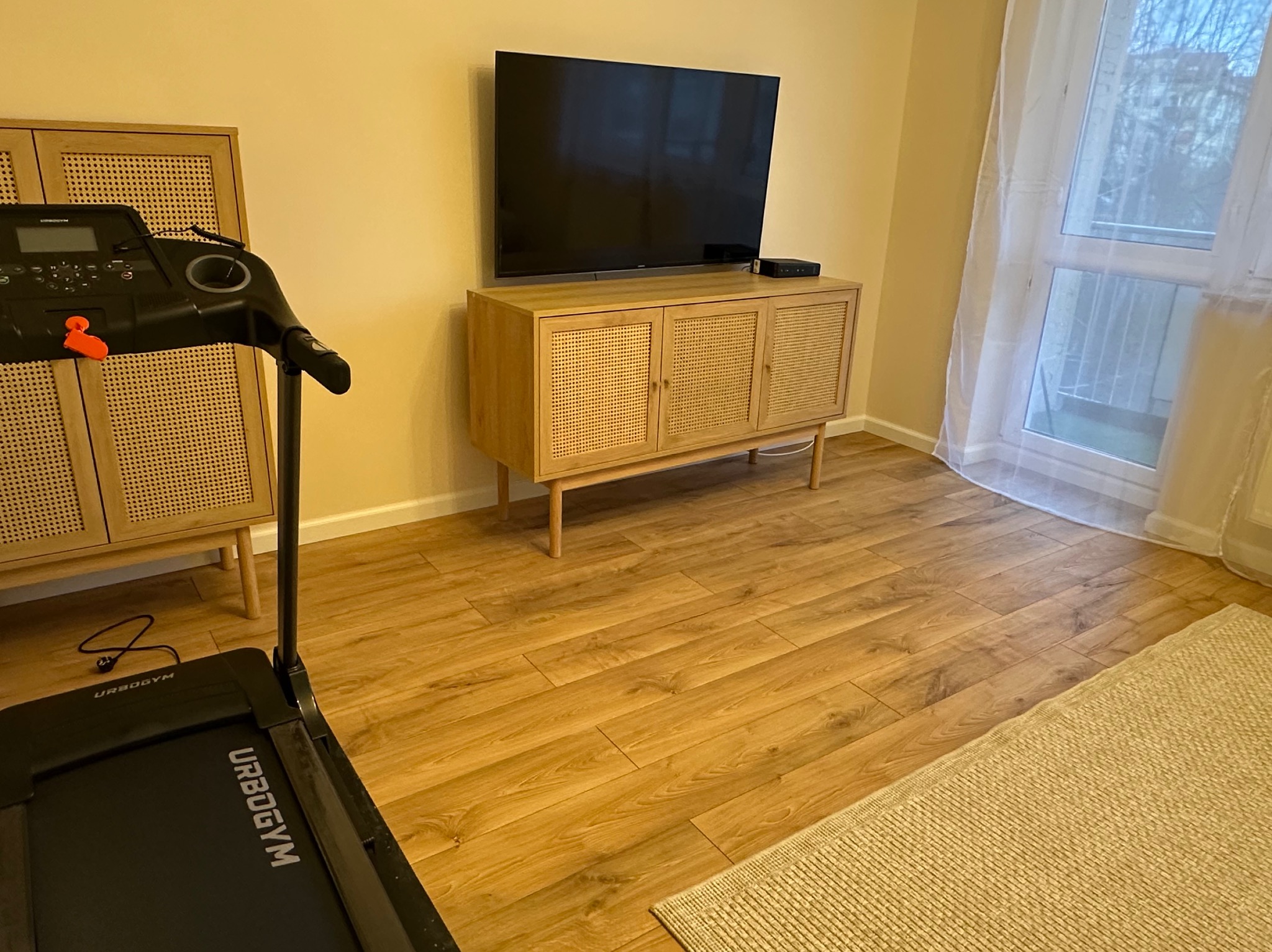 Jasne panele podłogowe w salonie z rattanową komodą pod telewizorem i bieżnią URBOGYM. Widok na okno z firanką i szafkę z rattanowymi frontami.