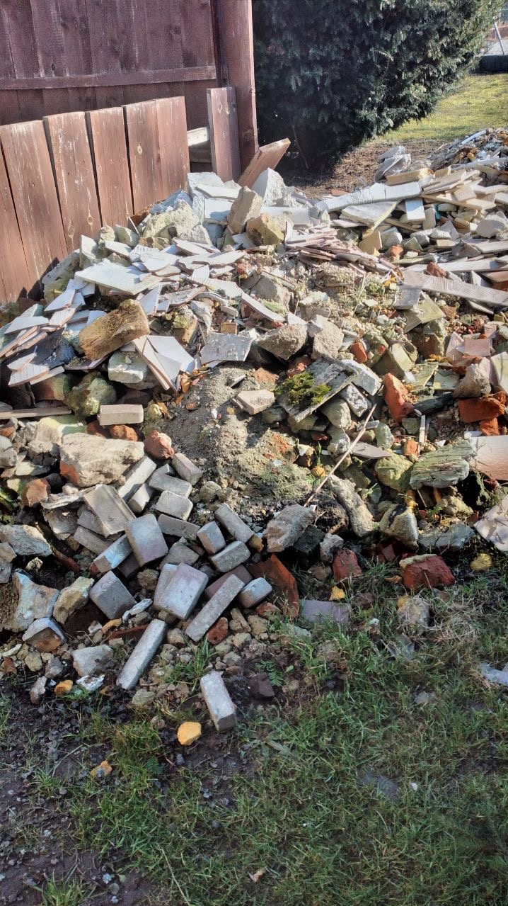 Kupa gruzu z cegieł, płytek i betonu leży na trawie obok drewnianego ogrodzenia. Fragment zieleni i drzewa w tle. Widoczne ślady wilgoci na elementach.