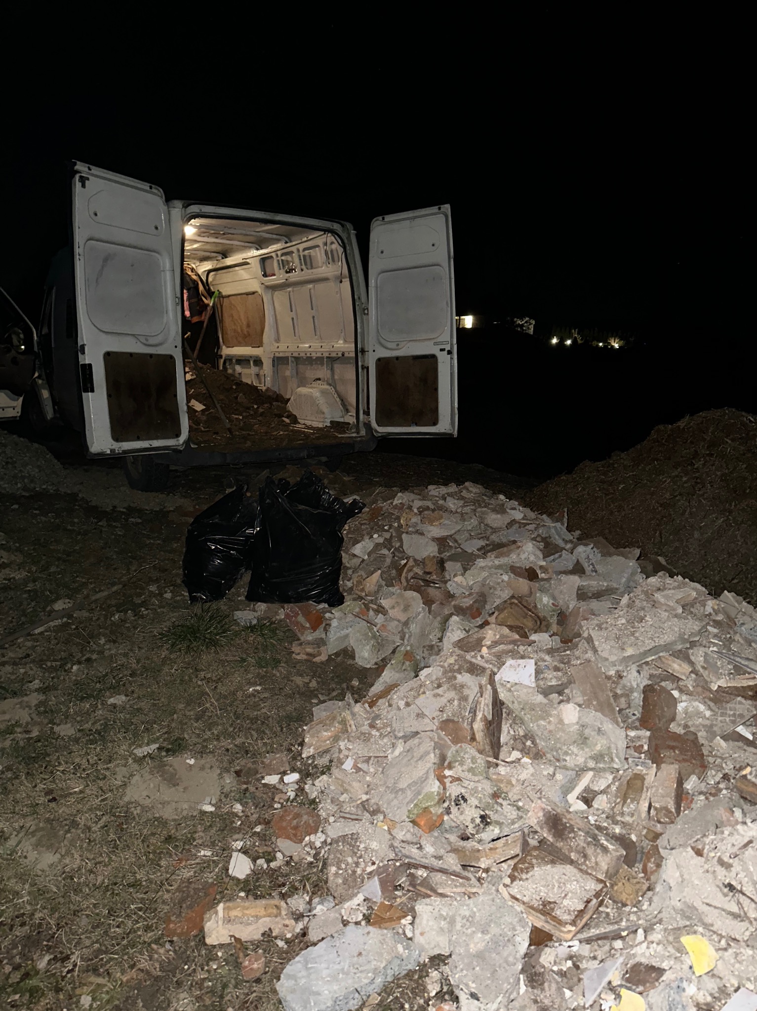 Nocne wywożenie gruzu busem: otwarte drzwi, wnętrze wypełnione odpadami, obok sterta gruzu i czarne worki. Słabe oświetlenie, widoczne elementy budynku w tle.