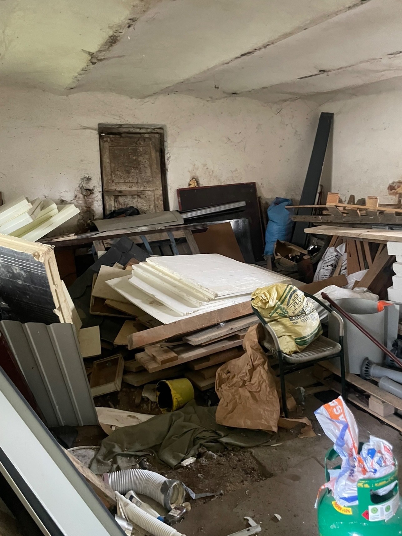 Zagracone pomieszczenie z różnymi przedmiotami, w tym styropianem, drewnem, workami i butlą gazową. Widoczne ślady wilgoci na ścianach i suficie. Nierówna podłoga.