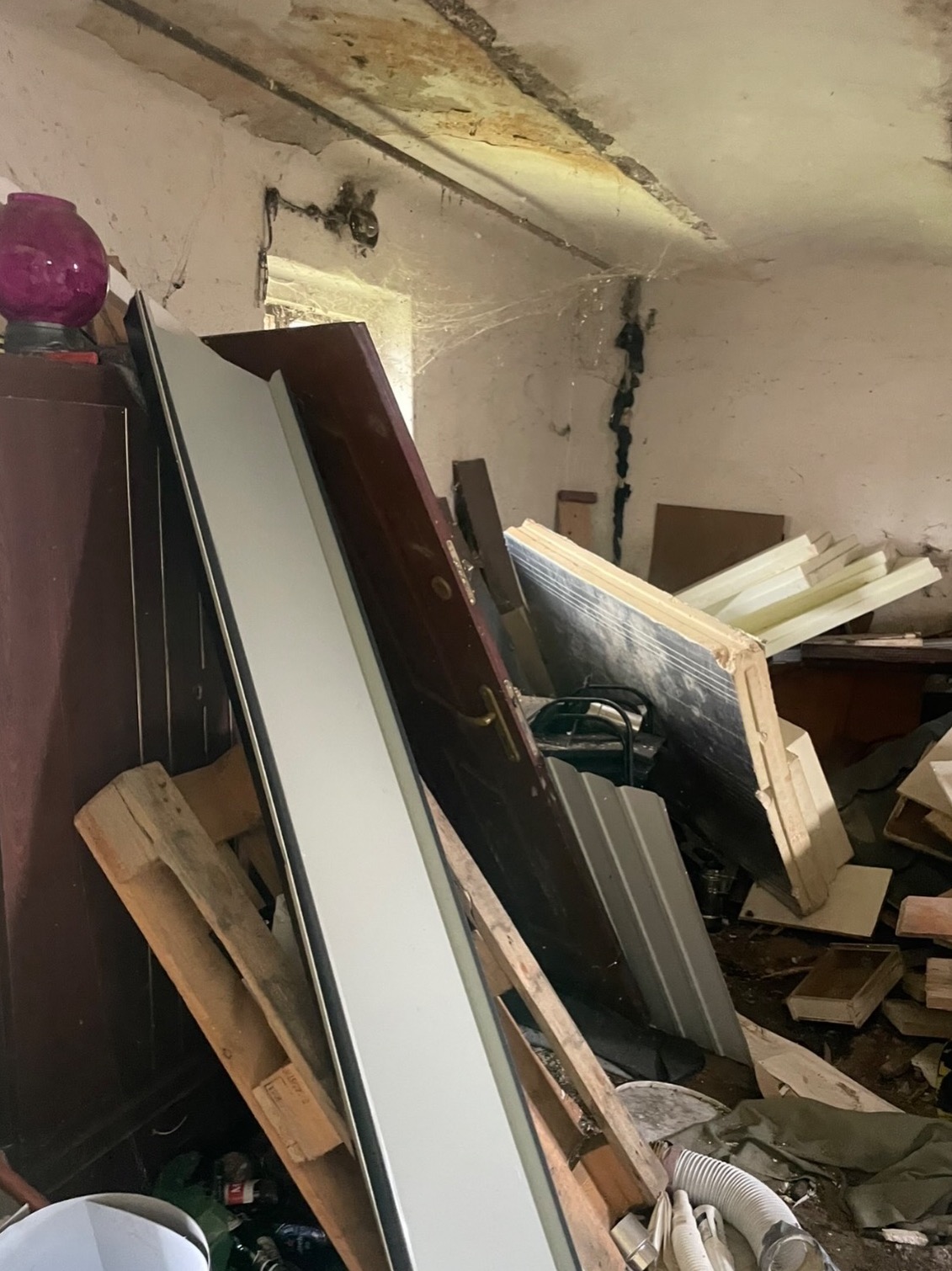 Stare drzwi, palety i gruz w zrujnowanym pomieszczeniu z pajęczynami. Widoczne ślady wilgoci na ścianach i suficie. Chaos i zaniedbanie.