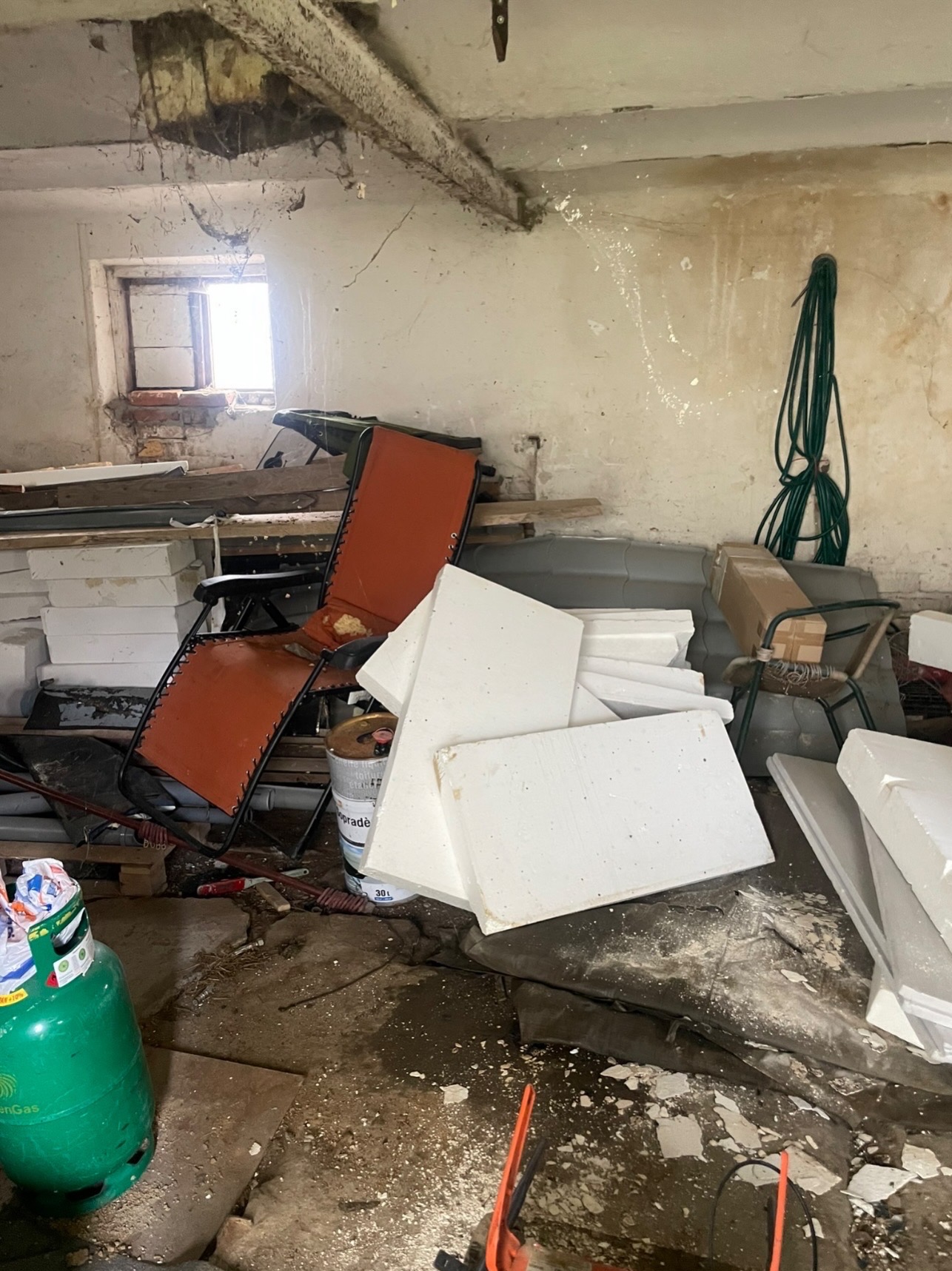 Zagracone pomieszczenie z oknem, wypełnione styropianem, starymi meblami i narzędziami. Widoczny bałagan i pozostałości po pracach budowlanych.