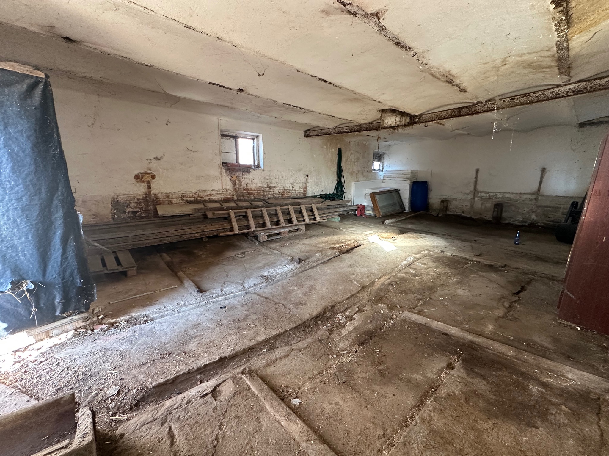 Wnętrze zaniedbanego pomieszczenia z widocznymi śladami wilgoci na ścianach i suficie. Na podłodze gruz, stare deski i drabina. Małe okno w tle.