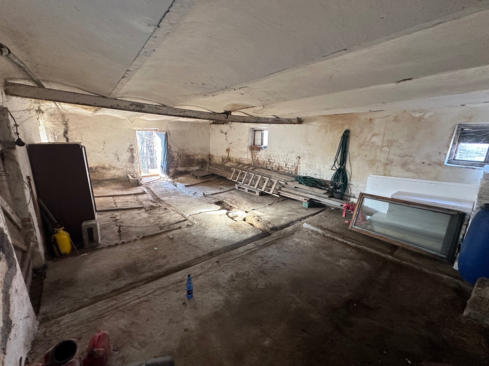 Wnętrze starego, zaniedbanego pomieszczenia z betonową podłogą, drabiną i oknem. Widoczne ślady wilgoci na ścianach i suficie. Otwór drzwiowy z widokiem na zewnątrz.