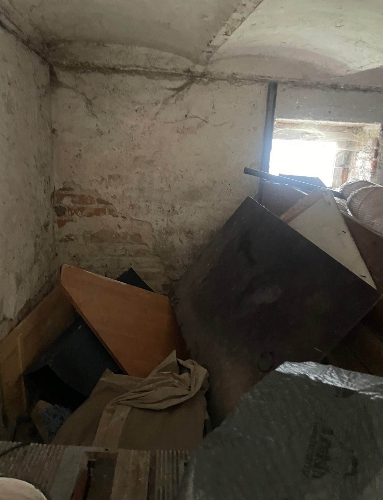 Zrujnowane wnętrze z gruzem i starymi meblami. Widoczne fragmenty cegieł i odpadający tynk na ścianach. W tle okno z jasnym światłem.
