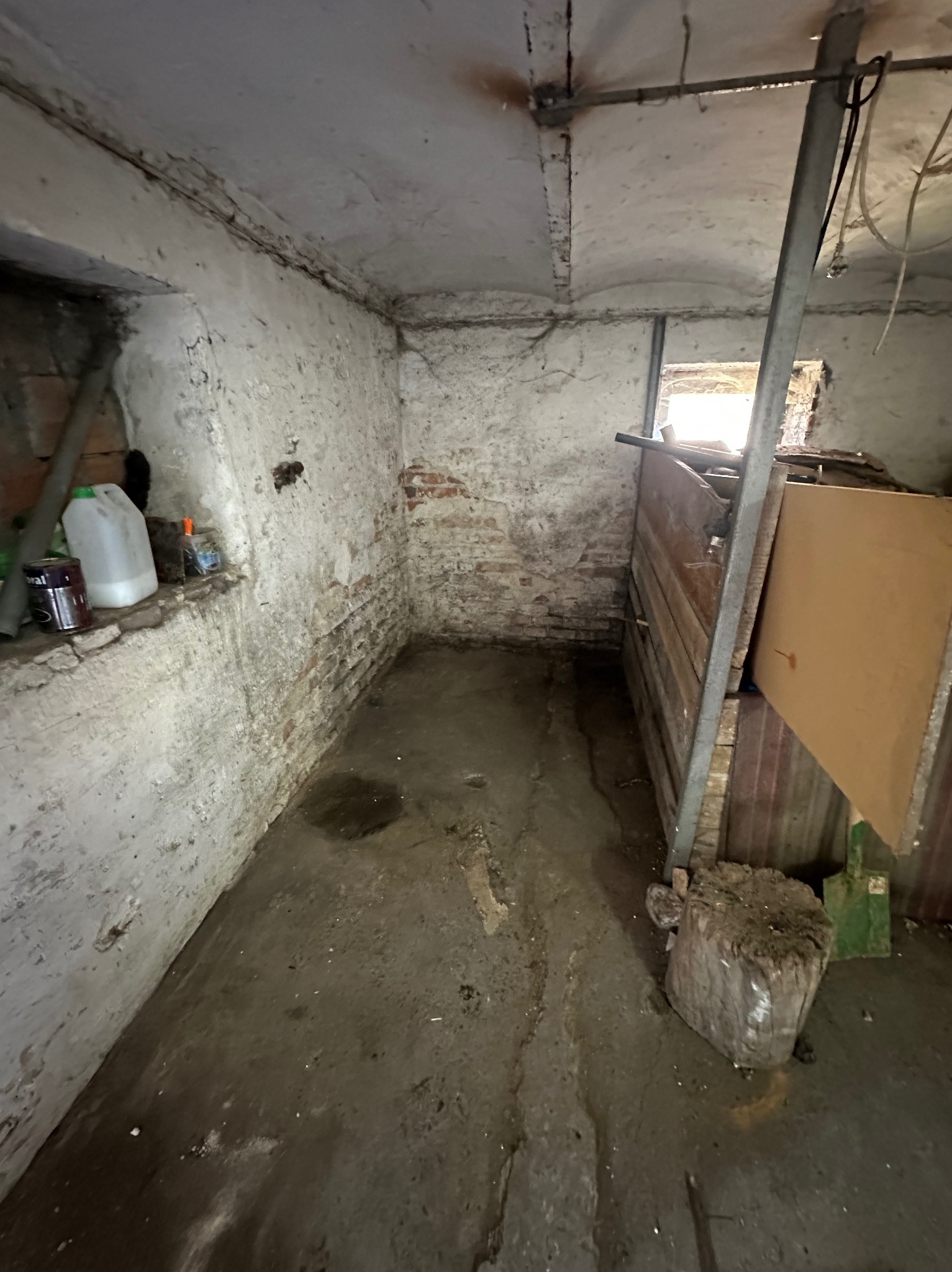 Zaniedbane, wilgotne pomieszczenie z oknem, widoczne ślady pleśni na ścianach, resztki przedmiotów i brudna podłoga. Nierówna faktura ścian i sufitu.