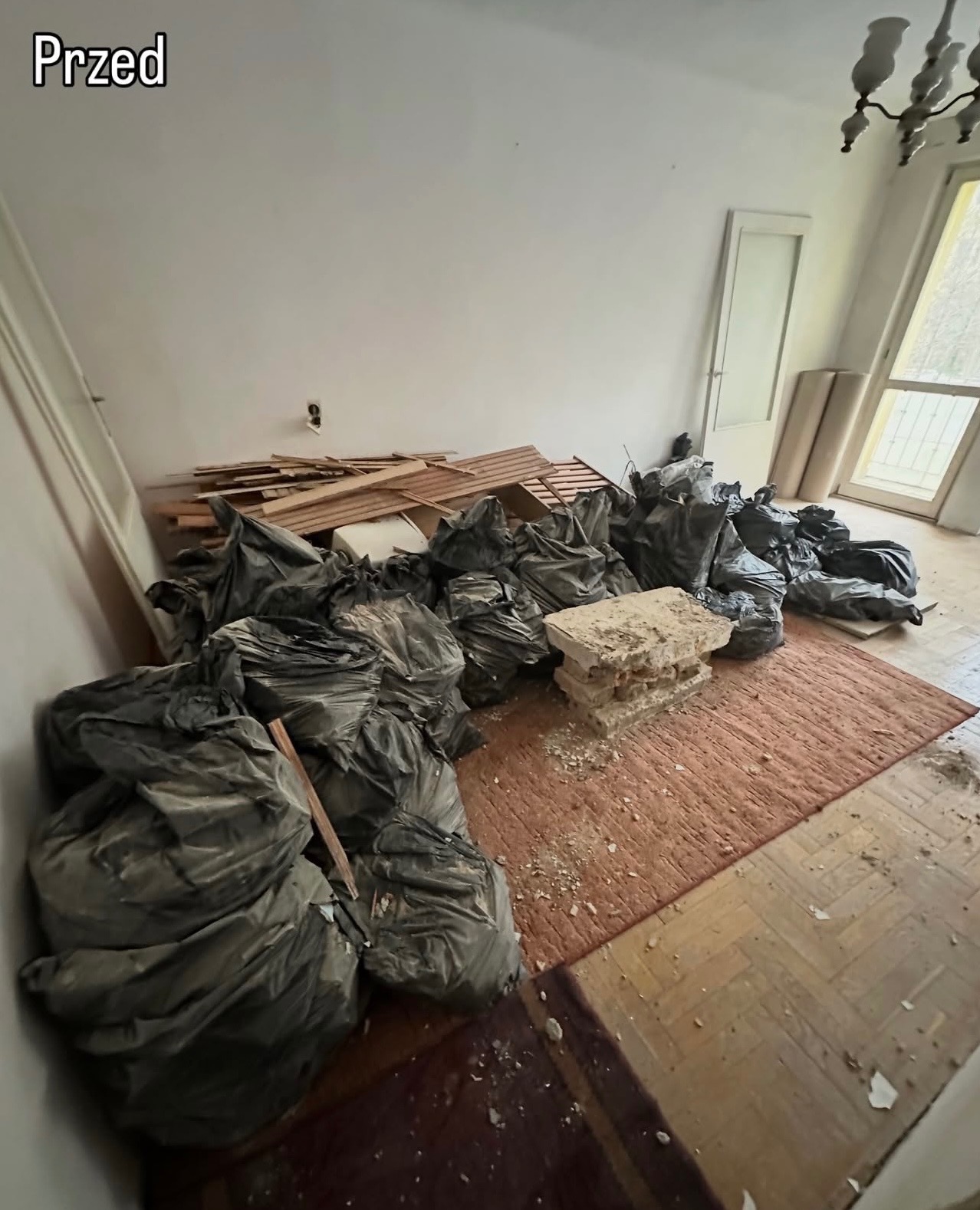 Pokój przed opróżnianiem: worki na śmieci, gruz, deski na dywanie i parkiecie. Widoczne okno i drzwi. Ujęcie z góry, oświetlenie naturalne.