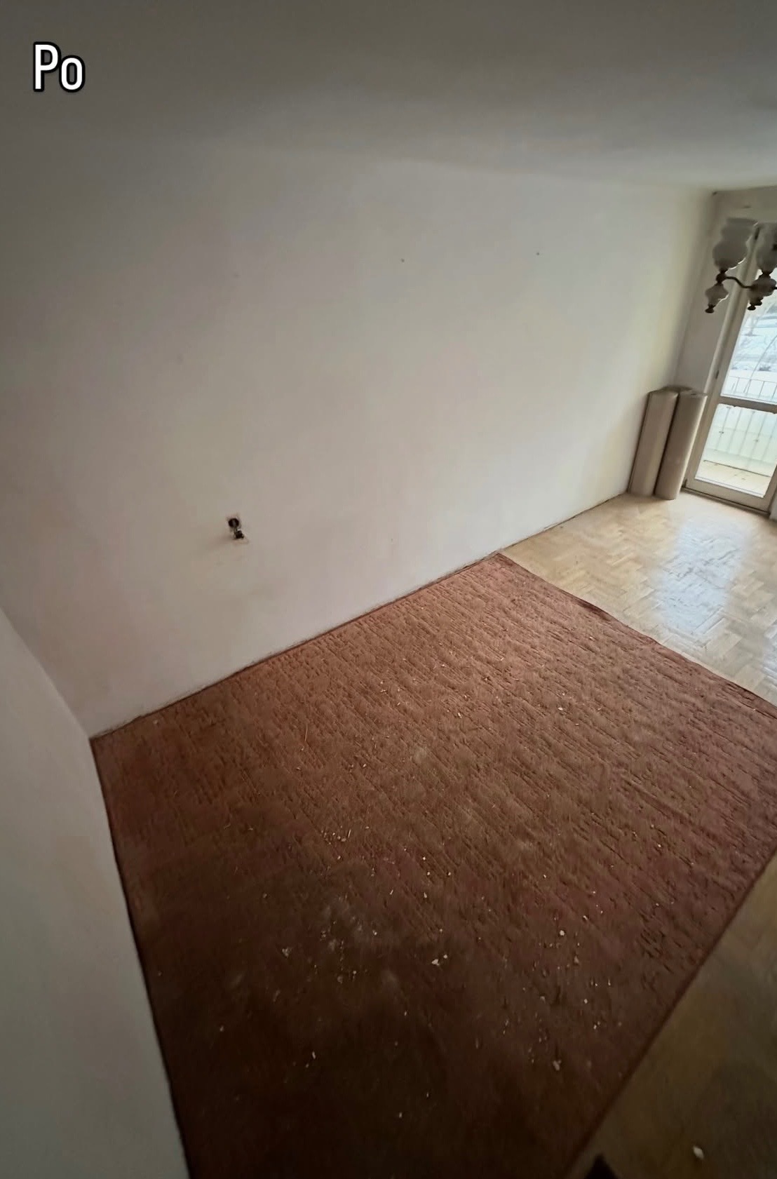 Pokój po remoncie: puste ściany, odnowiona podłoga z parkietu, częściowo zakryta dywanem. Widoczne ślady po gniazdku elektrycznym. Jasne wnętrze z oknem balkonowym.