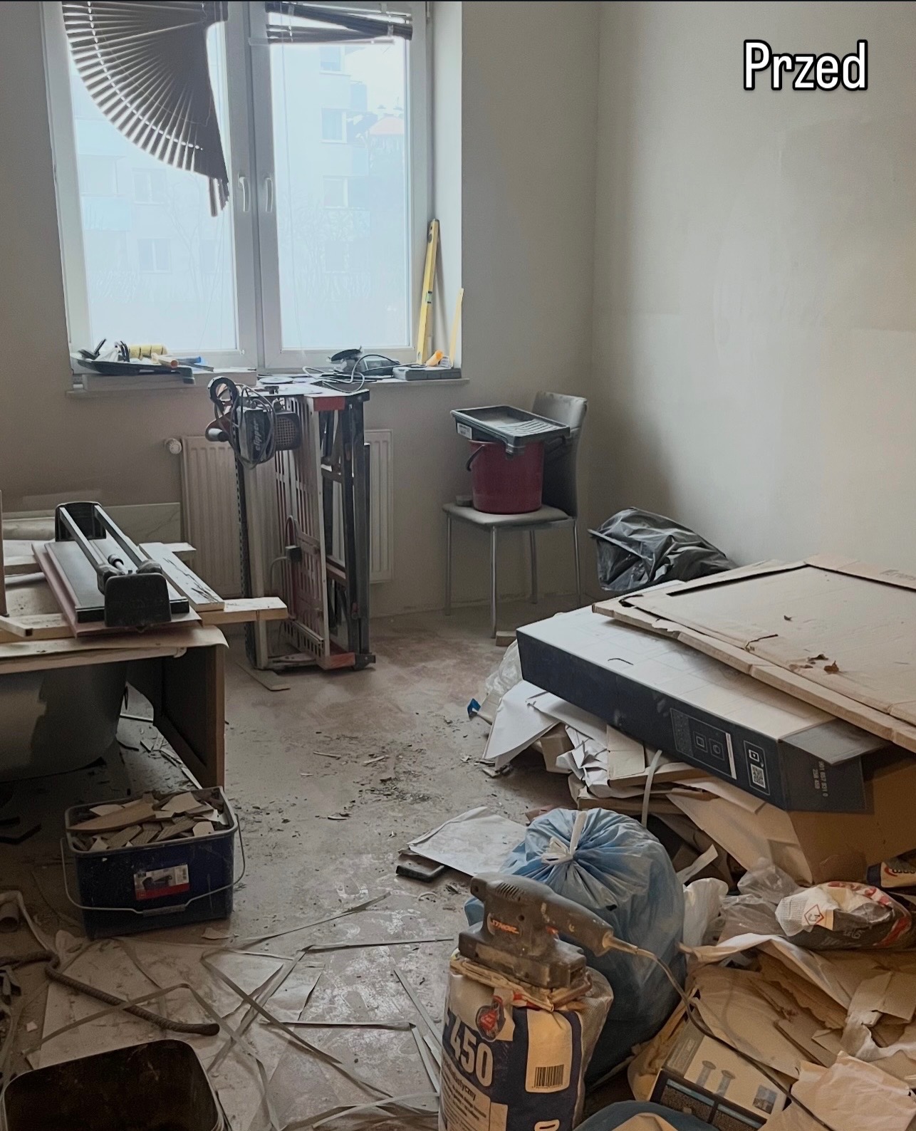 Stan 'Przed' remontem: wnętrze w Niechobrzu z narzędziami, resztkami materiałów i gruzem. Widoczna maszyna do cięcia płytek i worki z zaprawą.