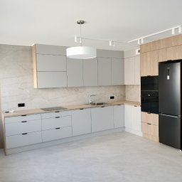 KERAD-MEBLE Dariusz Biliński - Jasna kuchnia z szarymi frontami i drewnianymi akcentami. Widoczna zabudowa kuchenna, sprzęt AGD w czarnym kolorze i marmurowa ściana. Nowoczesne oświetlenie sufitowe.