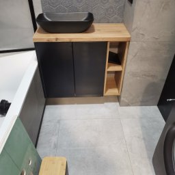 KERAD-MEBLE Dariusz Biliński - Nowoczesna szafka łazienkowa z czarnym blatem i umywalką, drewnianymi półkami. Szare płytki heksagonalne na ścianie. Widok z góry, fragment wanny i pralki.