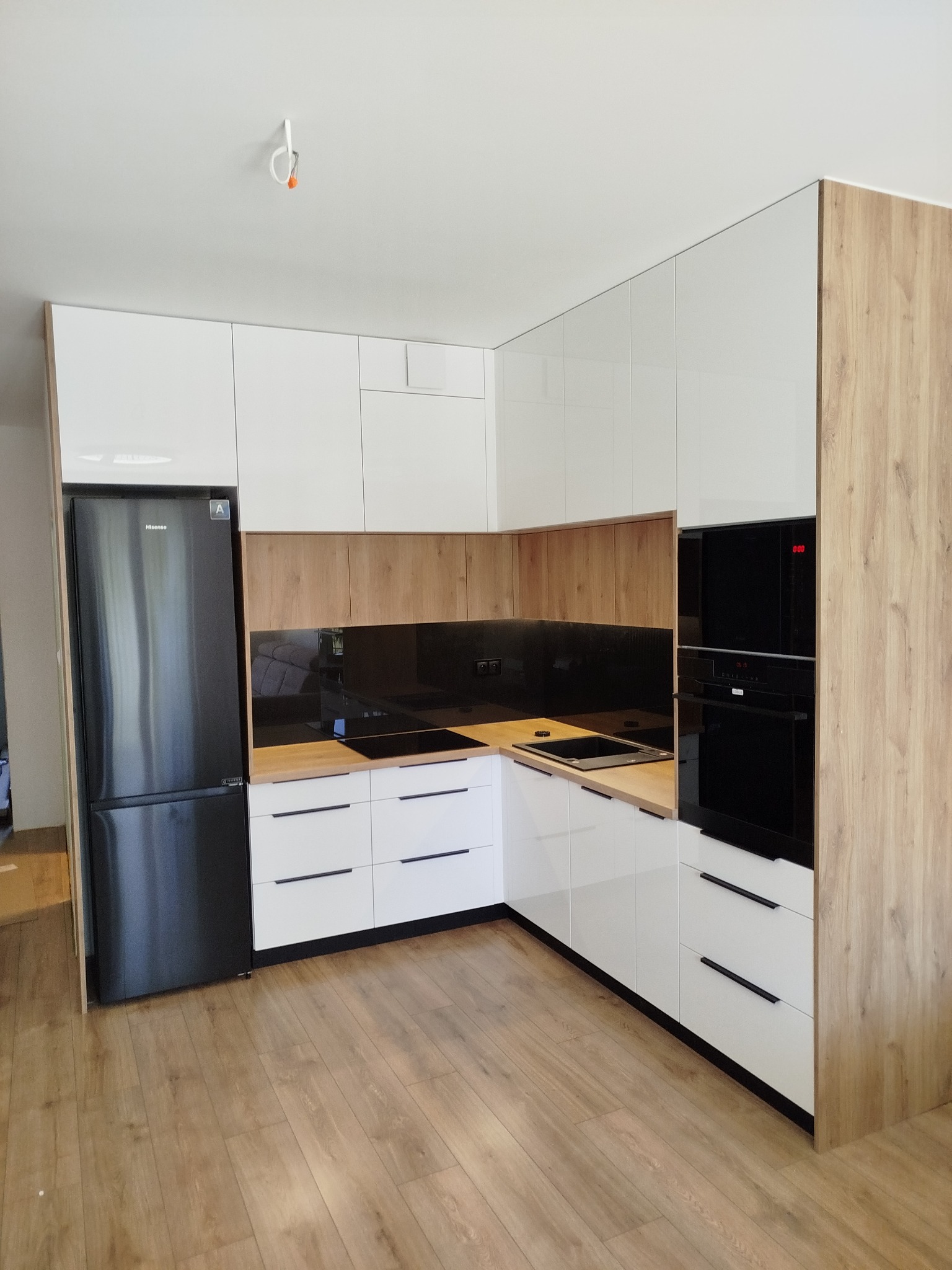 Nowoczesna kuchnia na wymiar: białe, lakierowane fronty szafek, drewnopodobne boki, czarny sprzęt AGD i blat z drewnianym dekorem. Minimalistyczny design.