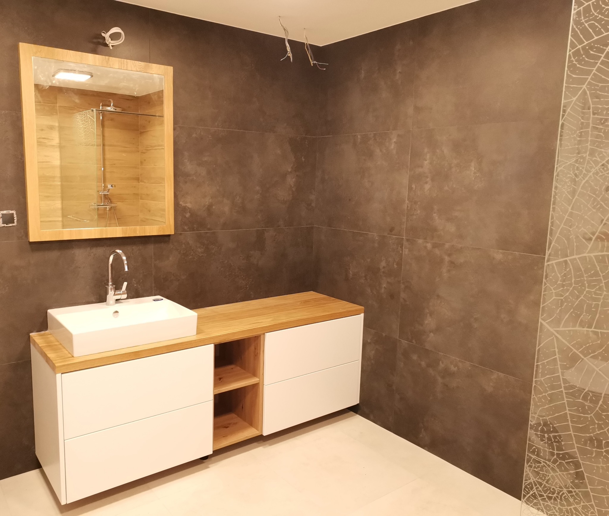 Nowoczesna łazienka z szafką pod umywalkę z drewnianym blatem i białymi frontami. Na ścianie lustro z widokiem na kabinę prysznicową. Ciemne płytki z motywem liści.