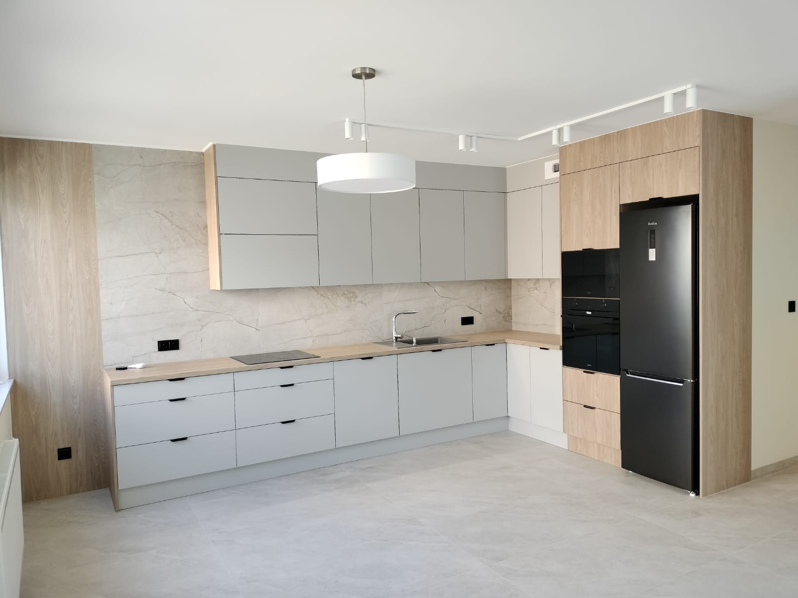 Jasna kuchnia z szarymi frontami i drewnianymi akcentami. Widoczna zabudowa kuchenna, sprzęt AGD w czarnym kolorze i marmurowa ściana. Nowoczesne oświetlenie sufitowe.
