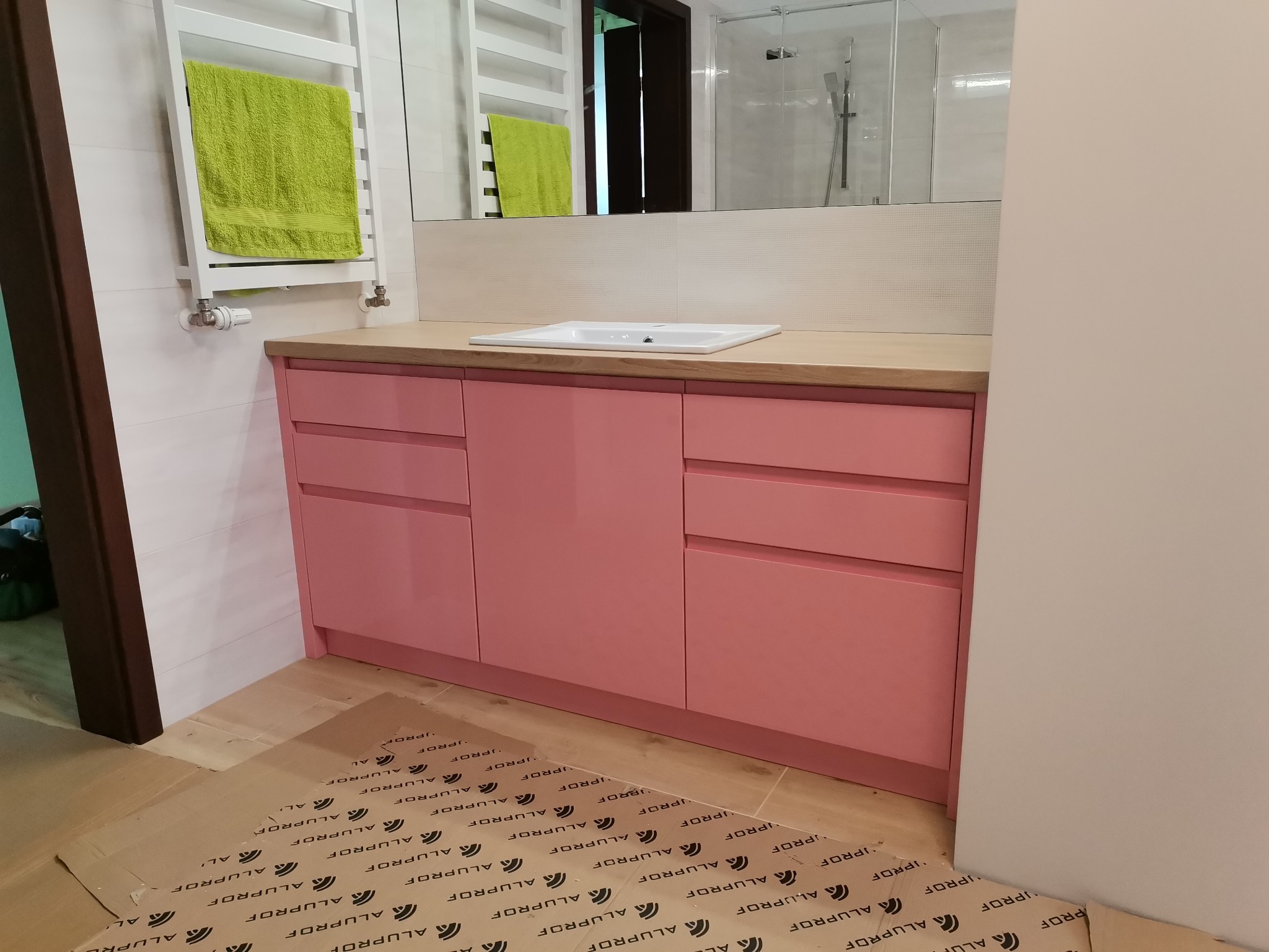 Różowa szafka łazienkowa z drewnianym blatem i białą umywalką w Olsztynie. Nad nią lustro, a obok grzejnik z zielonym ręcznikiem. Na podłodze folia ochronna.