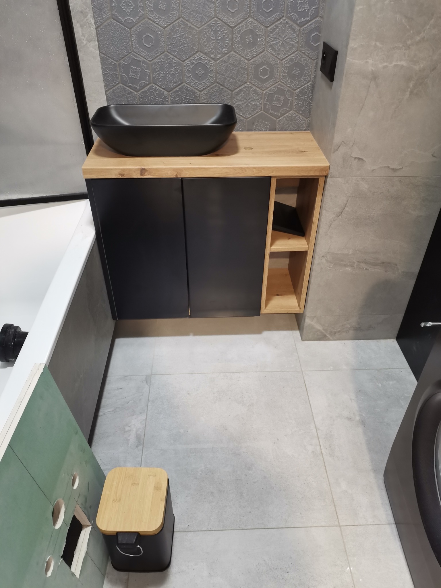 Nowoczesna szafka łazienkowa z czarnym blatem i umywalką, drewnianymi półkami. Szare płytki heksagonalne na ścianie. Widok z góry, fragment wanny i pralki.