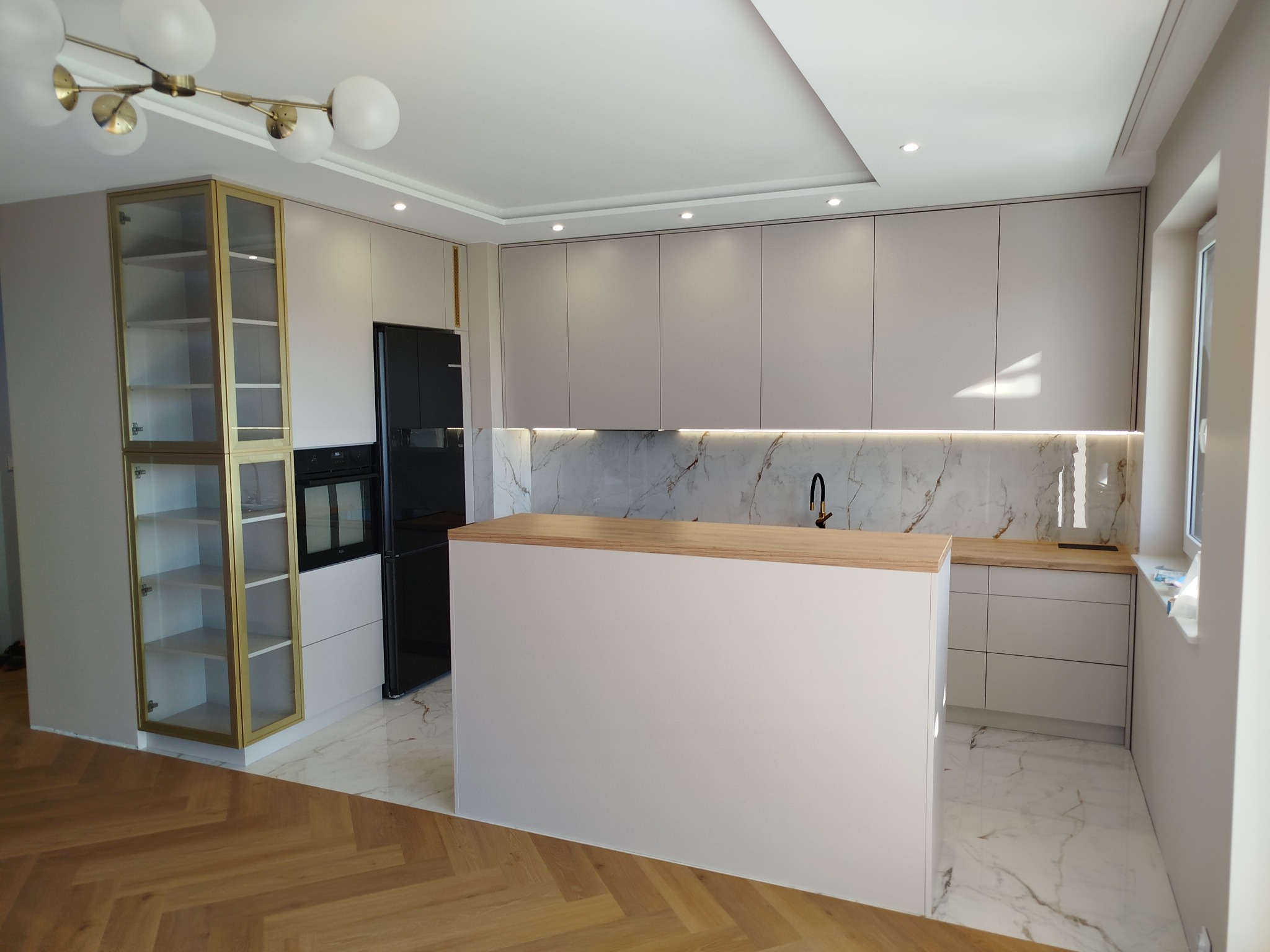 Jasna kuchnia z wyspą i złotymi akcentami: przeszklona witryna, marmurowy backsplash, drewniany blat. Nowoczesny design z oświetleniem LED.