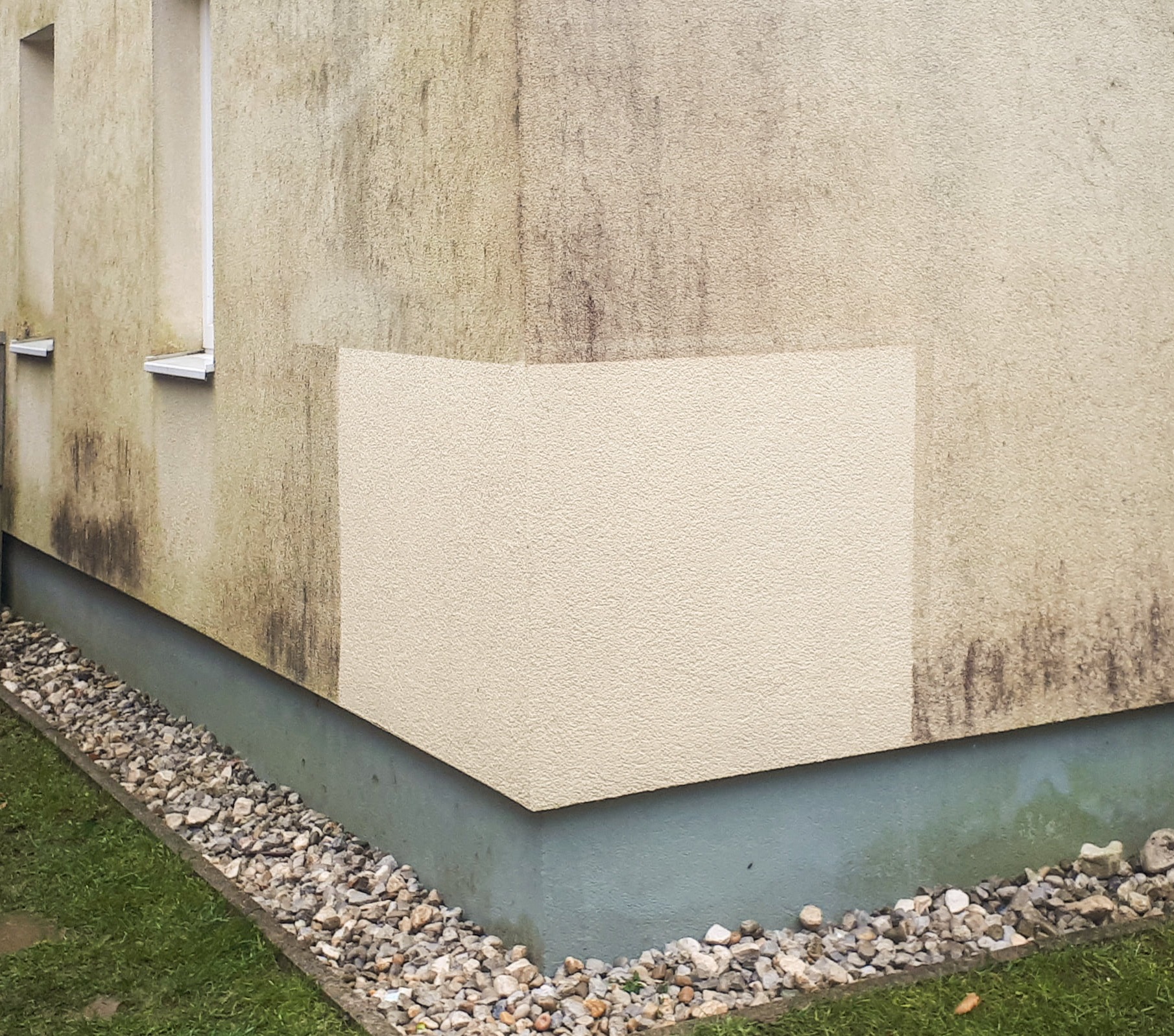 Fragment elewacji budynku z widocznym efektem czyszczenia: część ściany umyta, jaśniejsza, kontrastuje z brudną, ciemniejszą powierzchnią. Kamienie i trawa u podstawy.