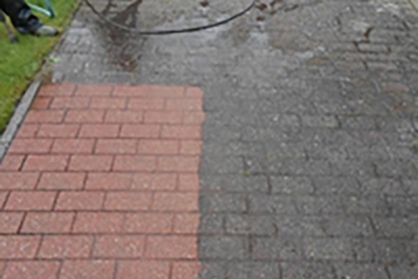 Częściowo wyczyszczona, czerwona kostka brukowa, z widoczną różnicą między czystą a brudną powierzchnią. W tle wąż i fragment buta osoby czyszczącej.