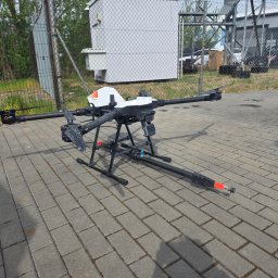 MJ Drone Sp. z o.o. - Dron ABZ Innovation C10 do mycia elewacji, dachów i przeszkleń, stojący na kostce brukowej. Widoczny przewód ciśnieniowy i dysza. W tle budynek i ogrodzenie.