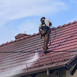 MJ Drone Sp. z o.o. - Pracownik w uprzęży bezpieczeństwa i kasku czyści czerwoną dachówkę myjką ciśnieniową, tworząc wyraźny strumień wody. Widoczne są fragmenty dachu przed i po myciu.