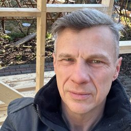 Leszek Dulinski - Konstrukcja drewniana w trakcie budowy, widoczna przez ramię mężczyzny w kurtce. W tle zieleń ogrodu i elementy drewnianej konstrukcji.