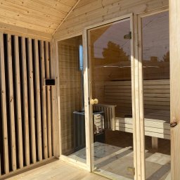 Leszek Dulinski - Jasna, drewniana sauna z przeszklonymi drzwiami, widok z zewnątrz. Widoczne drewniane ławki wewnątrz i piec. Ściana z pionowych listew po lewej stronie.