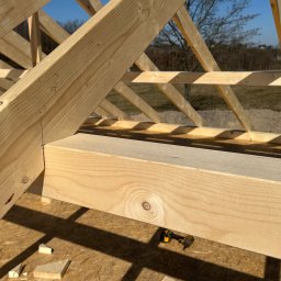 Leszek Dulinski - Konstrukcja drewniana dachu w trakcie budowy, widoczne elementy więźby i narzędzie pracy. Jasne drewno, słoneczny dzień.
