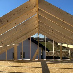 Leszek Dulinski - Konstrukcja drewniana dachu w trakcie budowy, widok od wewnątrz. Jasne drewno na tle błękitnego nieba i fragmentu budynku w oddali. Widoczne elementy złączne.