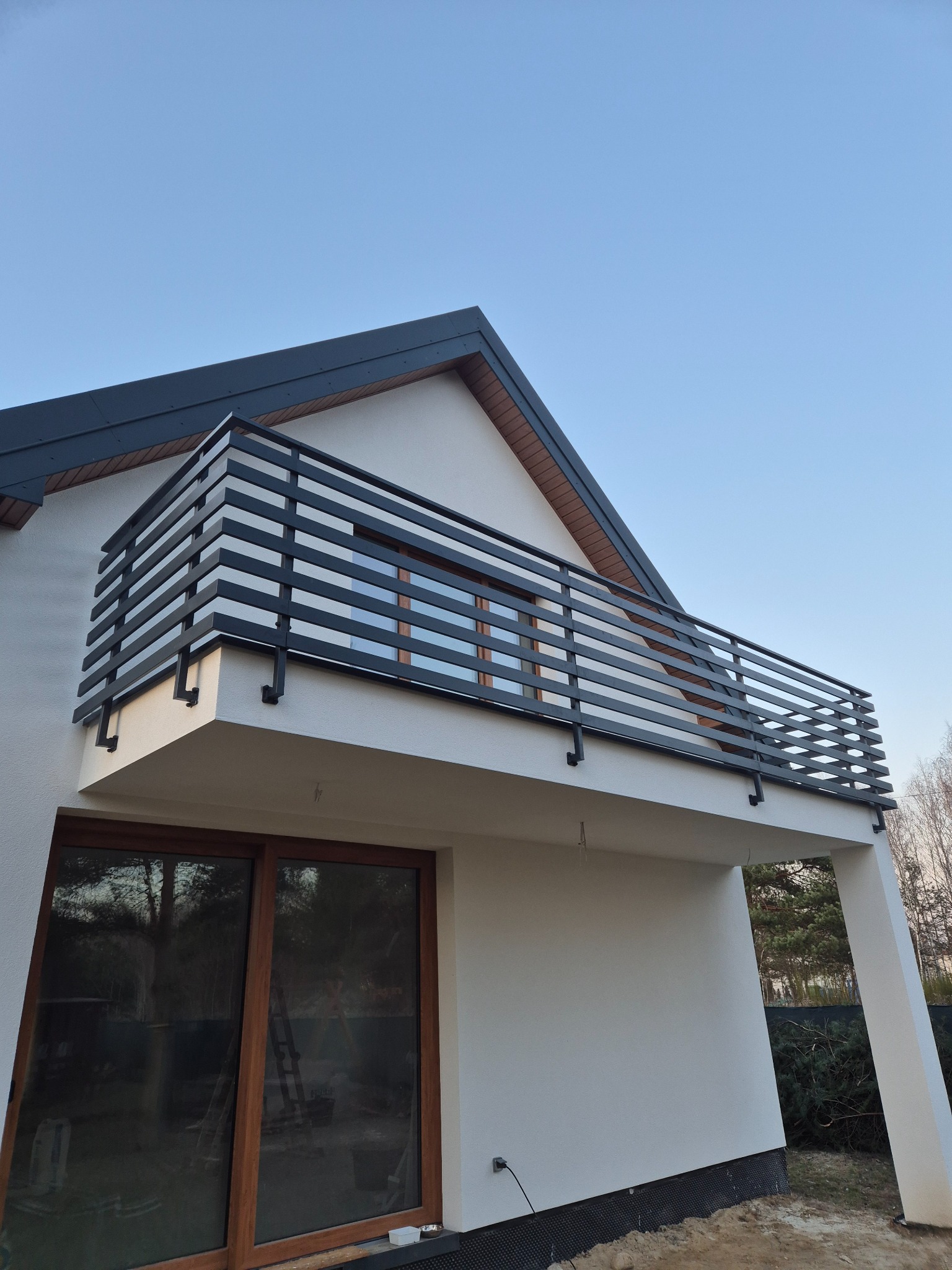 Nowoczesna, czarna balustrada balkonowa z poziomych profili, zamontowana na białym budynku. Minimalistyczny design, widok z dołu, błękitne niebo w tle.