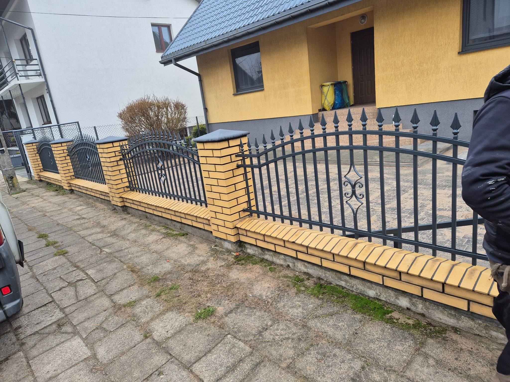 Ciemna, metalowa konstrukcja ogrodzenia z dekoracyjnymi elementami, osadzona na niskim murku z żółtej cegły klinkierowej, na tle domu z żółtą elewacją.