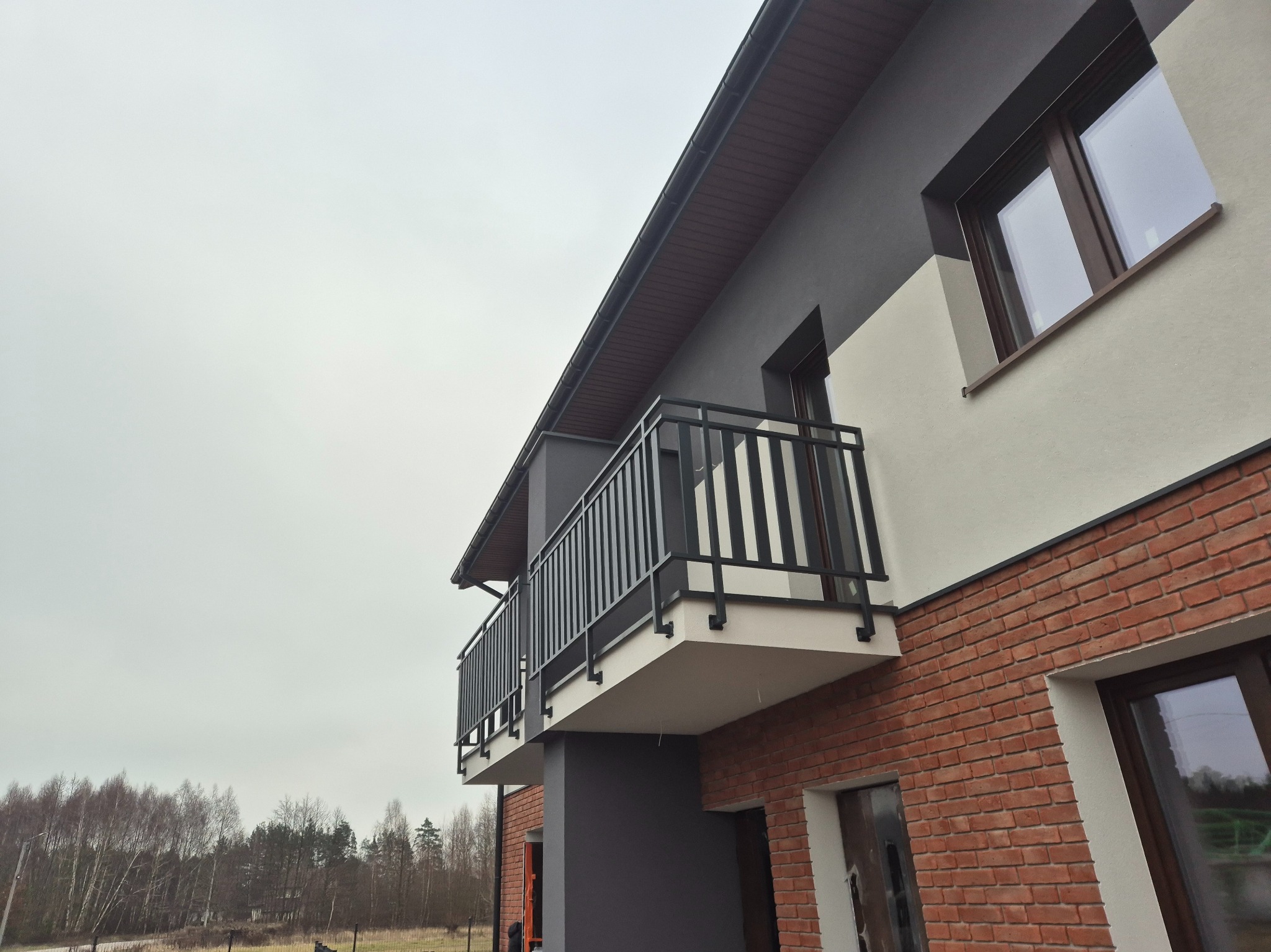 Nowoczesna, czarna balustrada balkonowa z pionowymi szczeblami, zamontowana w Augustowie. Widok z dołu, na tle ceglanej elewacji i pochmurnego nieba.