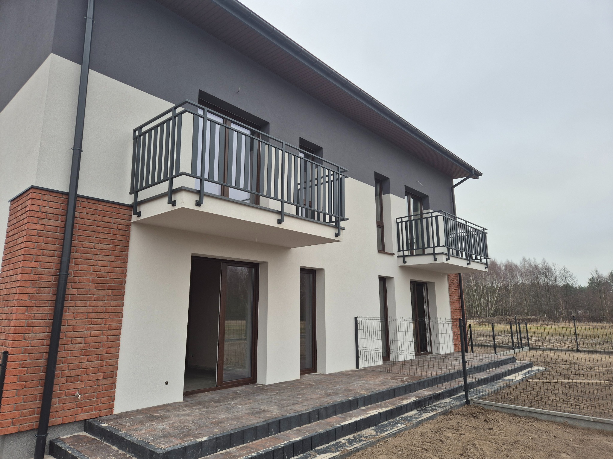 Nowoczesny dom z balkonami z czarnymi barierkami i ogrodzeniem panelowym. Widok z zewnątrz na elewację z ceglanym akcentem i schodami.