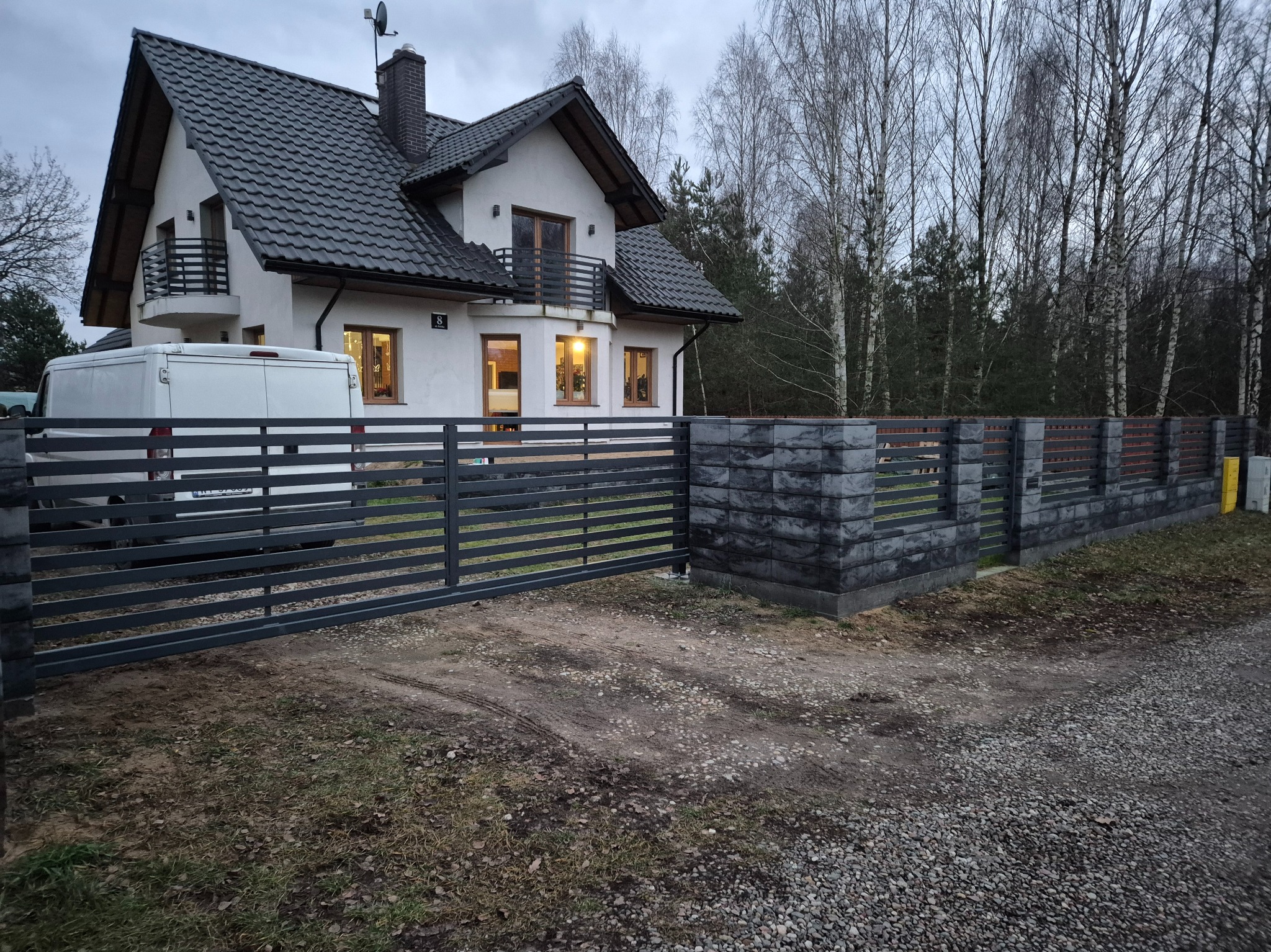 Nowoczesne ogrodzenie panelowe z elementami imitującymi kamień, na tle domu z balkonem i lasu. Szara kolorystyka, poziome panele. Widoczny fragment białego samochodu.