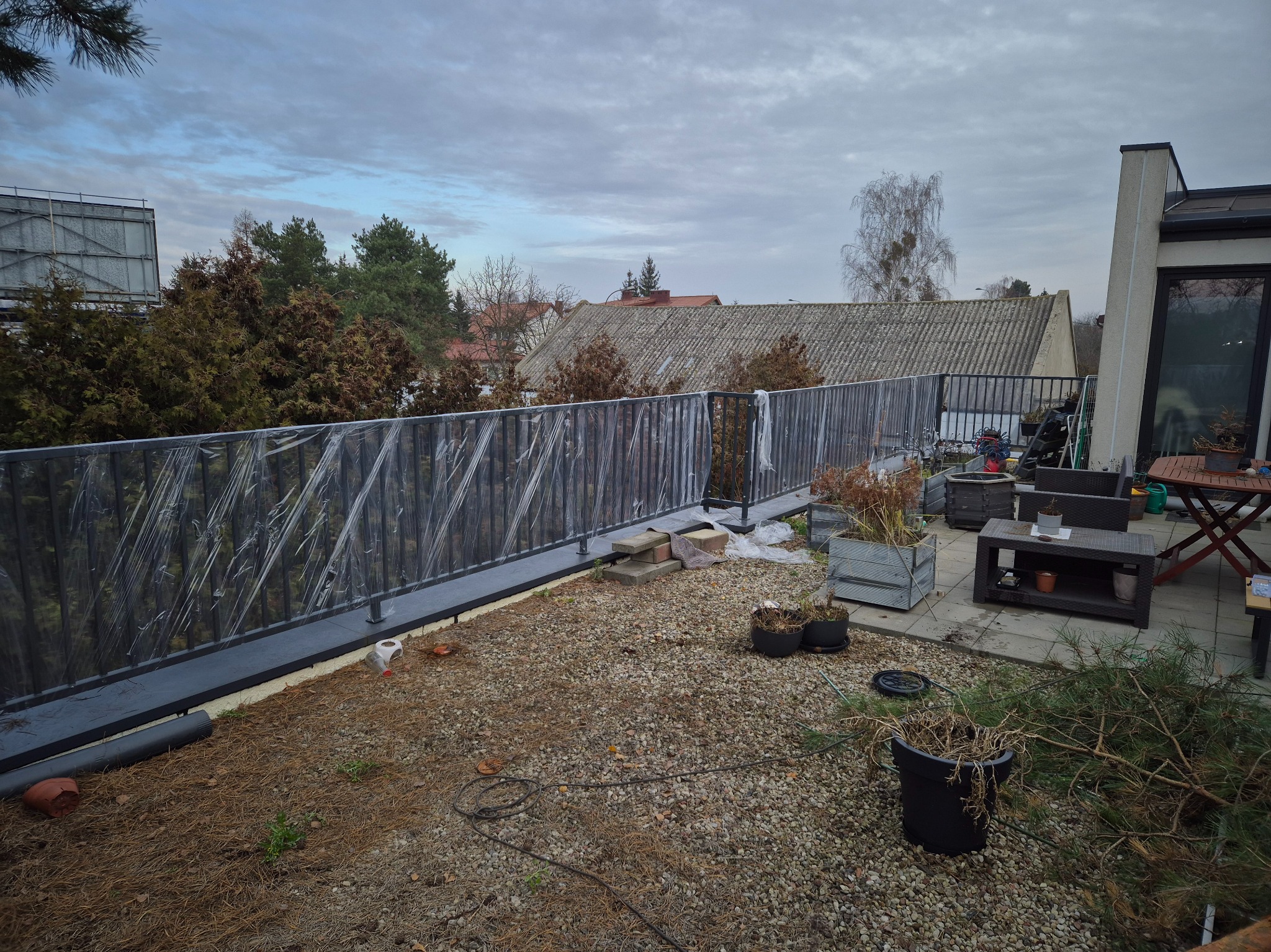 Szara, metalowa barierka z folią ochronną na balkonie z kamienną nawierzchnią. W tle budynki i drzewa pod pochmurnym niebem. Widok z góry.