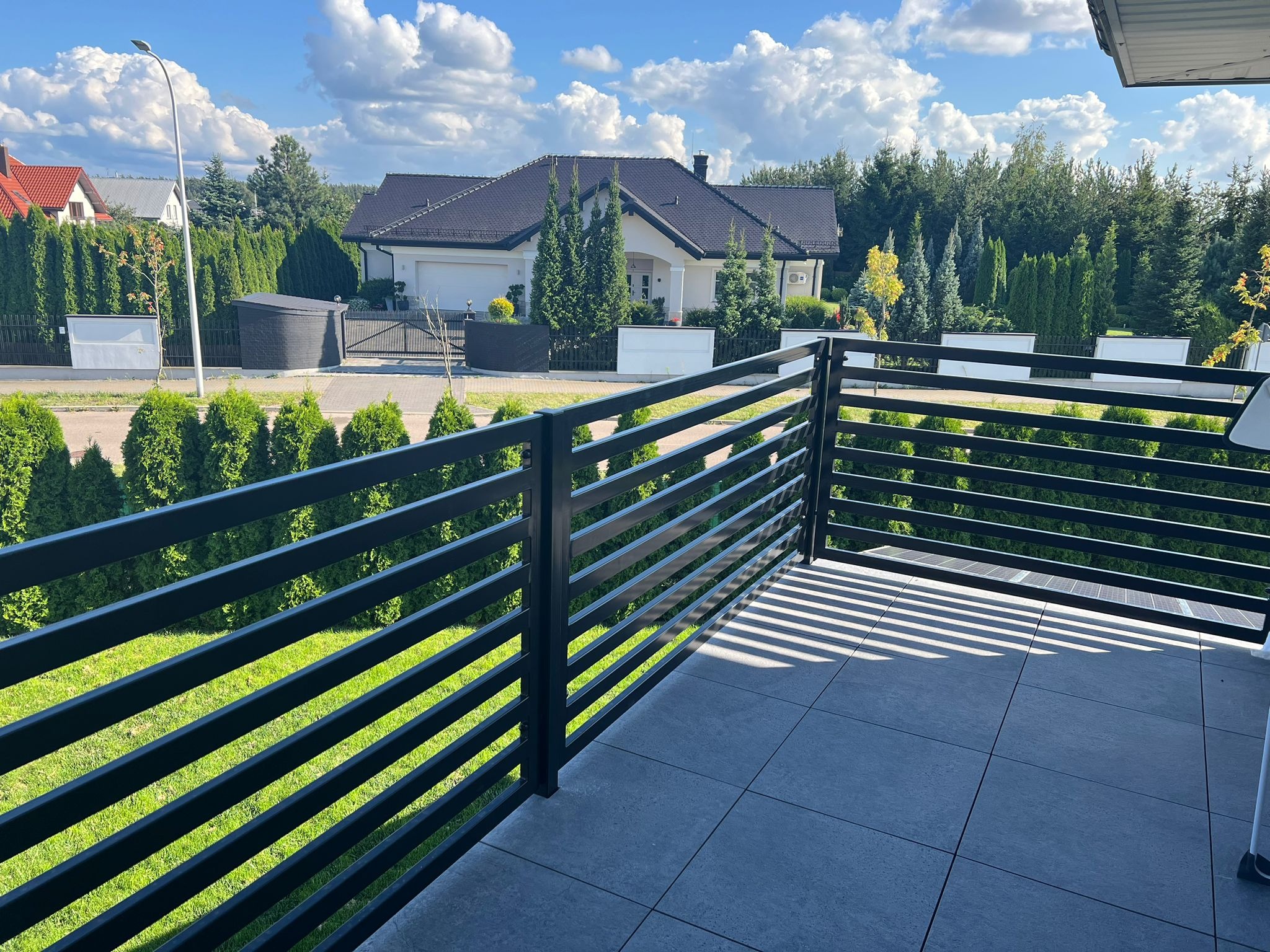 Nowoczesna, czarna balustrada balkonowa z poziomych profili na tle zadbanego ogrodu i domu jednorodzinnego w słoneczny dzień. Widoczne cienie rzucane przez konstrukcję.