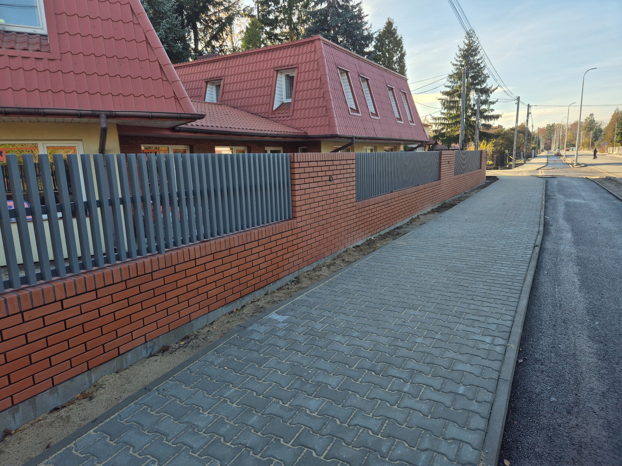 Nowoczesne ogrodzenie z szarych, pionowych paneli i ceglanej podmurówki, oddzielające posesję od chodnika. W tle dom z czerwoną dachówką i drzewa.