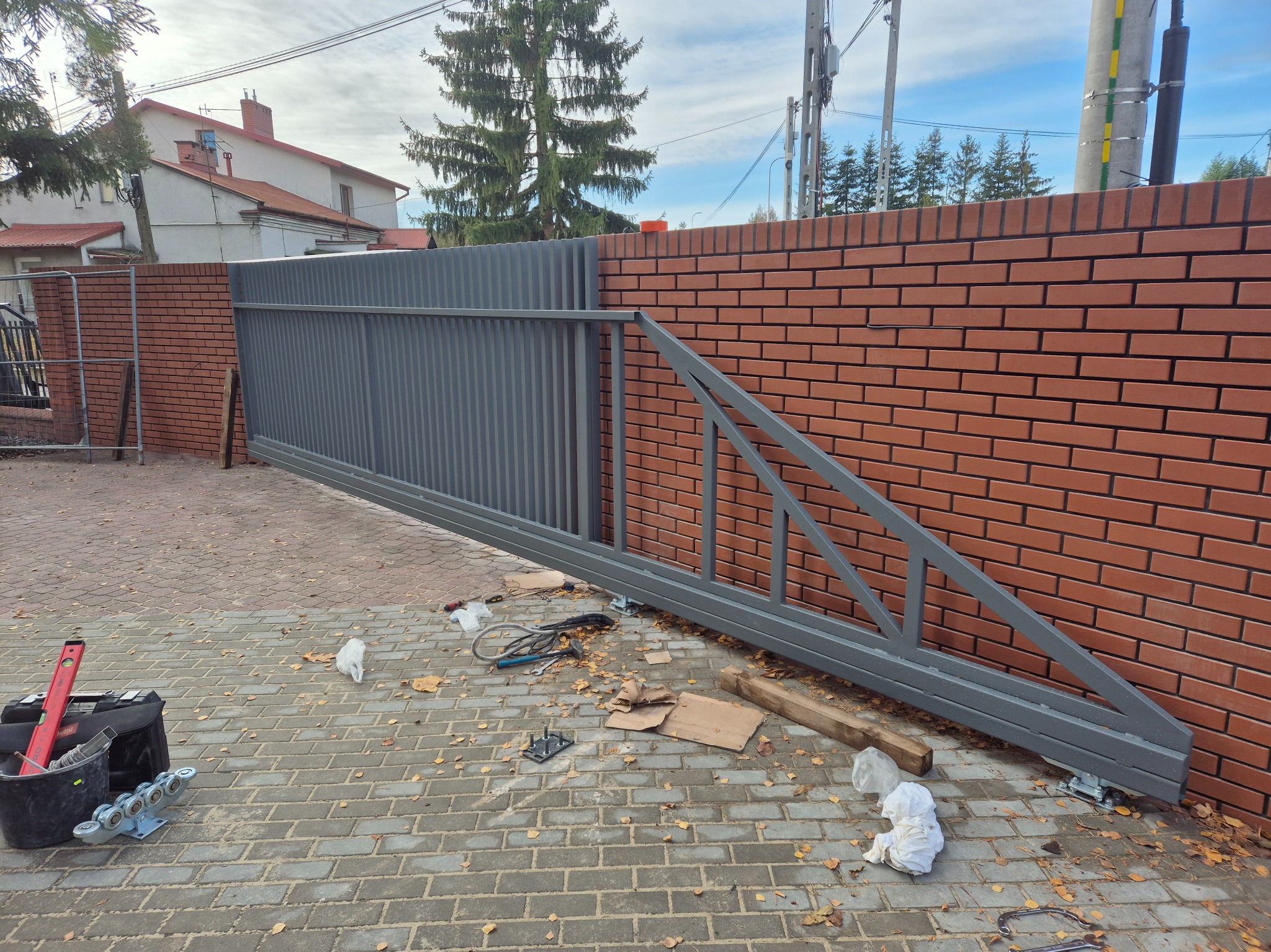 Montaż przesuwnej bramy wjazdowej z szarego metalu, widoczne narzędzia i akcesoria na kostce brukowej. Brama przylega do muru z czerwonej cegły.