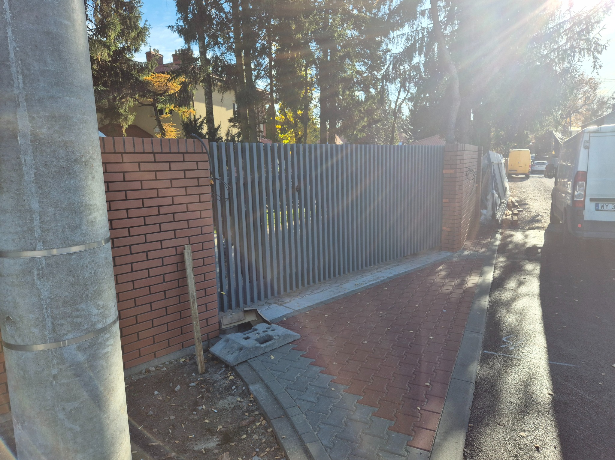 Nowoczesne ogrodzenie posesji z pionowych, szarych paneli metalowych, osadzone na ceglanych słupkach. Widoczny fragment chodnika z kostki i słup energetyczny.