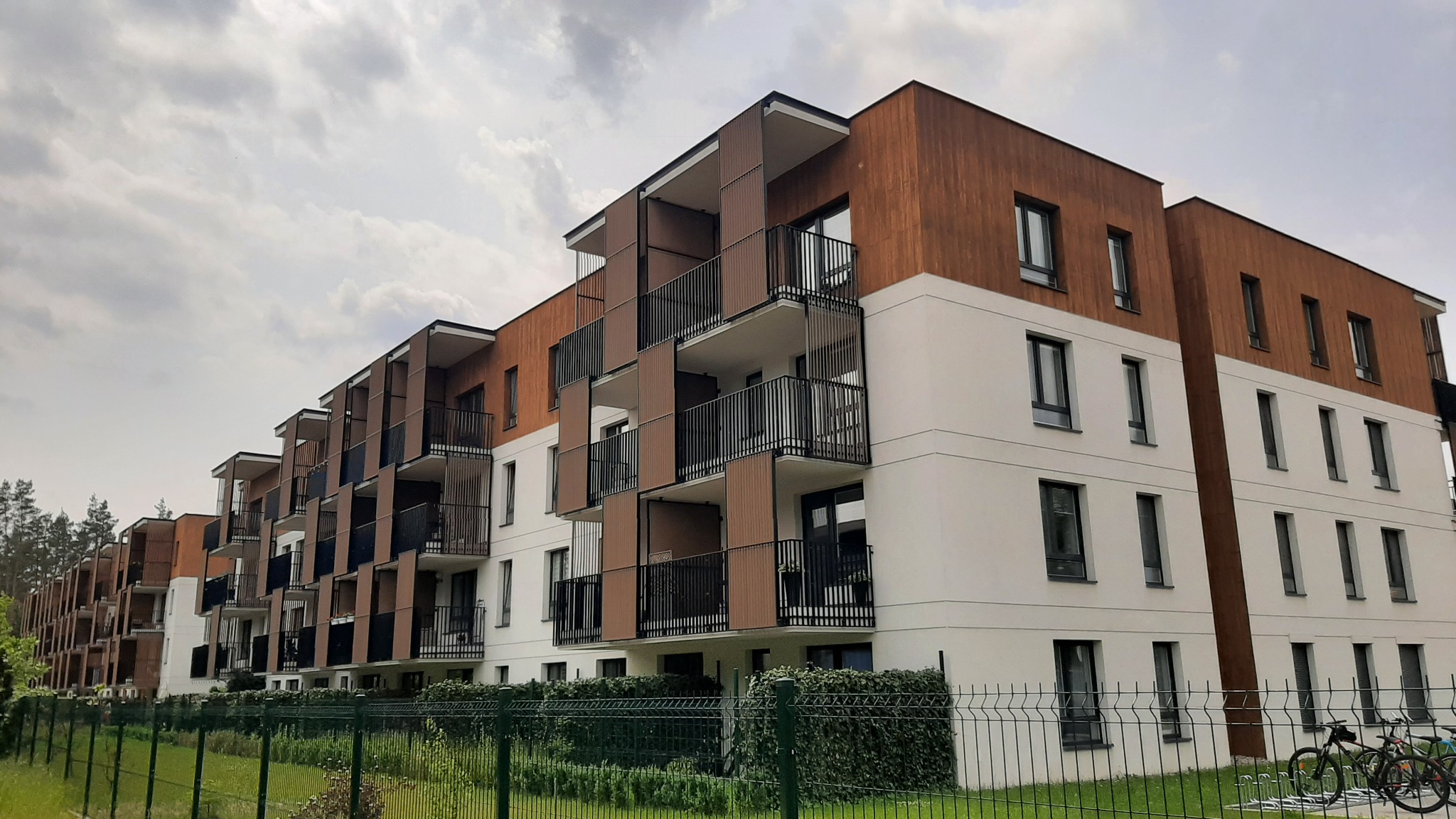 Nowoczesny budynek wielorodzinny z balkonami zabezpieczonymi czarnymi, metalowymi barierkami. W tle widoczne drzewa i pochmurne niebo.