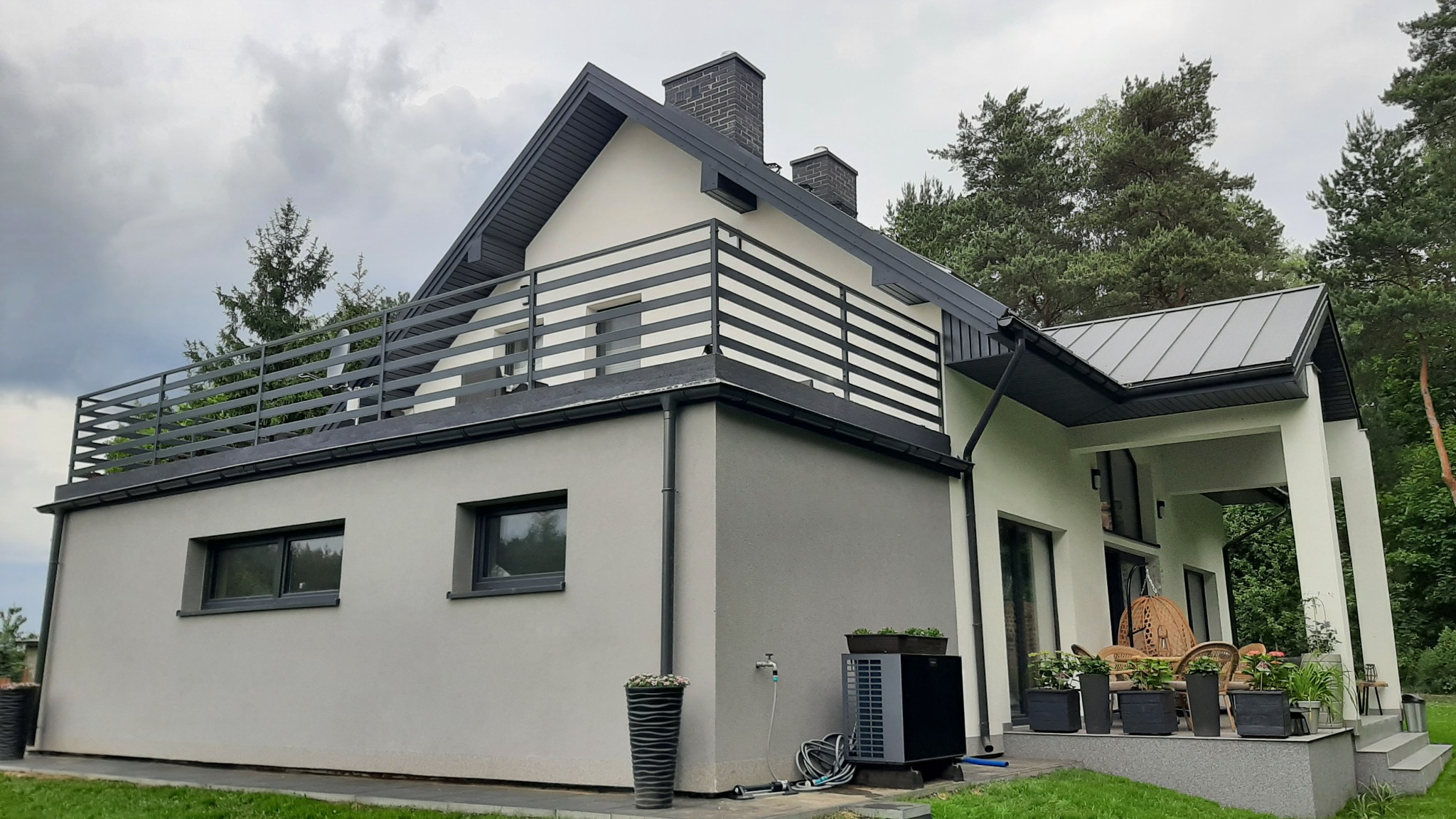 Nowoczesny dom z balkonem zabezpieczonym poziomą, metalową konstrukcją. Szara kolorystyka budynku i balustrady, widok z boku, zieleń w tle.