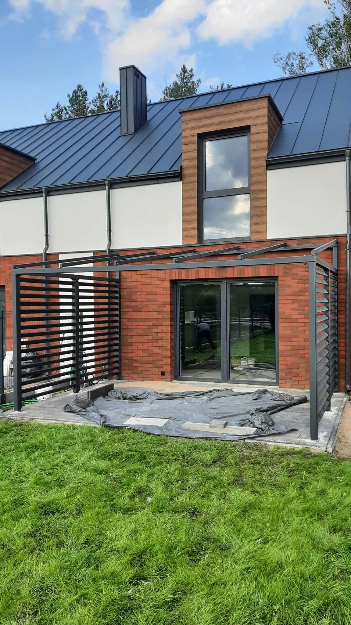 Nowoczesna pergola z metalowymi lamelami przy domu z ceglaną elewacją. Widoczne przygotowania do dalszych prac na tarasie.