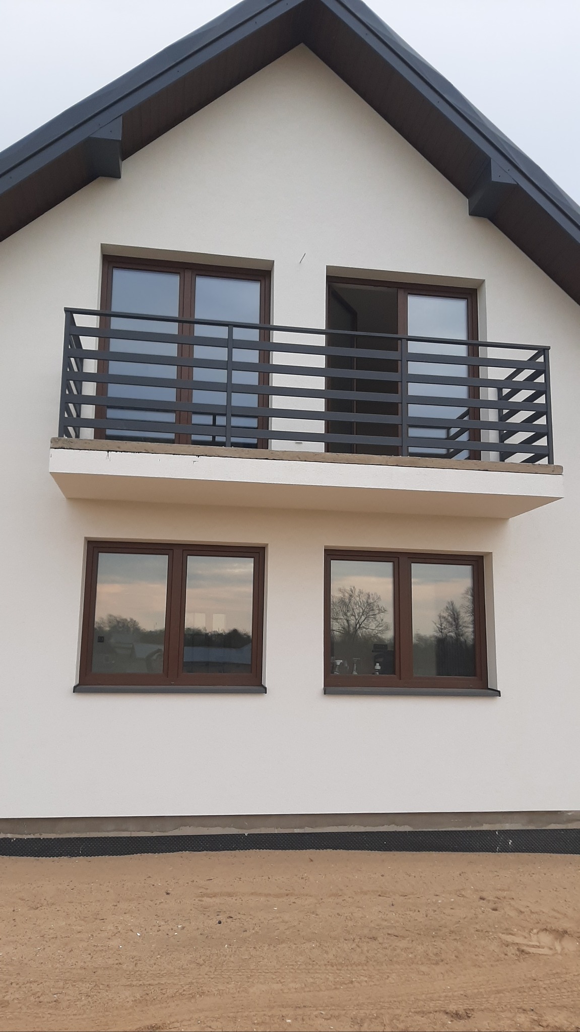 Nowoczesna, pozioma balustrada balkonowa z ciemnego metalu na tle białej elewacji domu z brązowymi oknami. Widok z dołu, surowy teren wokół budynku.