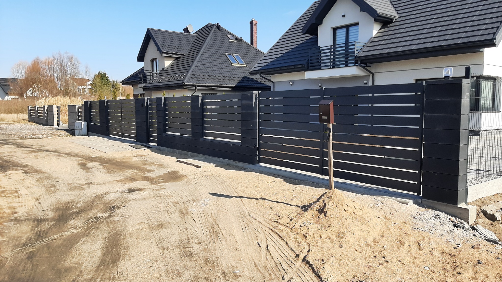 Nowoczesne, poziome ogrodzenie metalowe w kolorze antracytowym, z bramą wjazdową i furtką, na tle domu jednorodzinnego w słoneczny dzień. Widoczny fragment podjazdu.