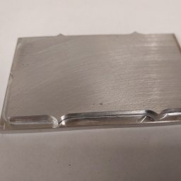 Edgecut Łukasz Chudzik - Szlifowany, prostokątny element metalowy z precyzyjnie wyfrezowanymi wgłębieniami na krawędziach, leżący na jasnym tle. Widoczne ślady obróbki.