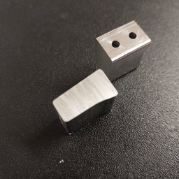 Edgecut Łukasz Chudzik - Dwa precyzyjnie obrobione, metalowe prostopadłościany z otworami na ciemnym tle. Jeden leży, drugi stoi, ukazując detale obróbki CNC.