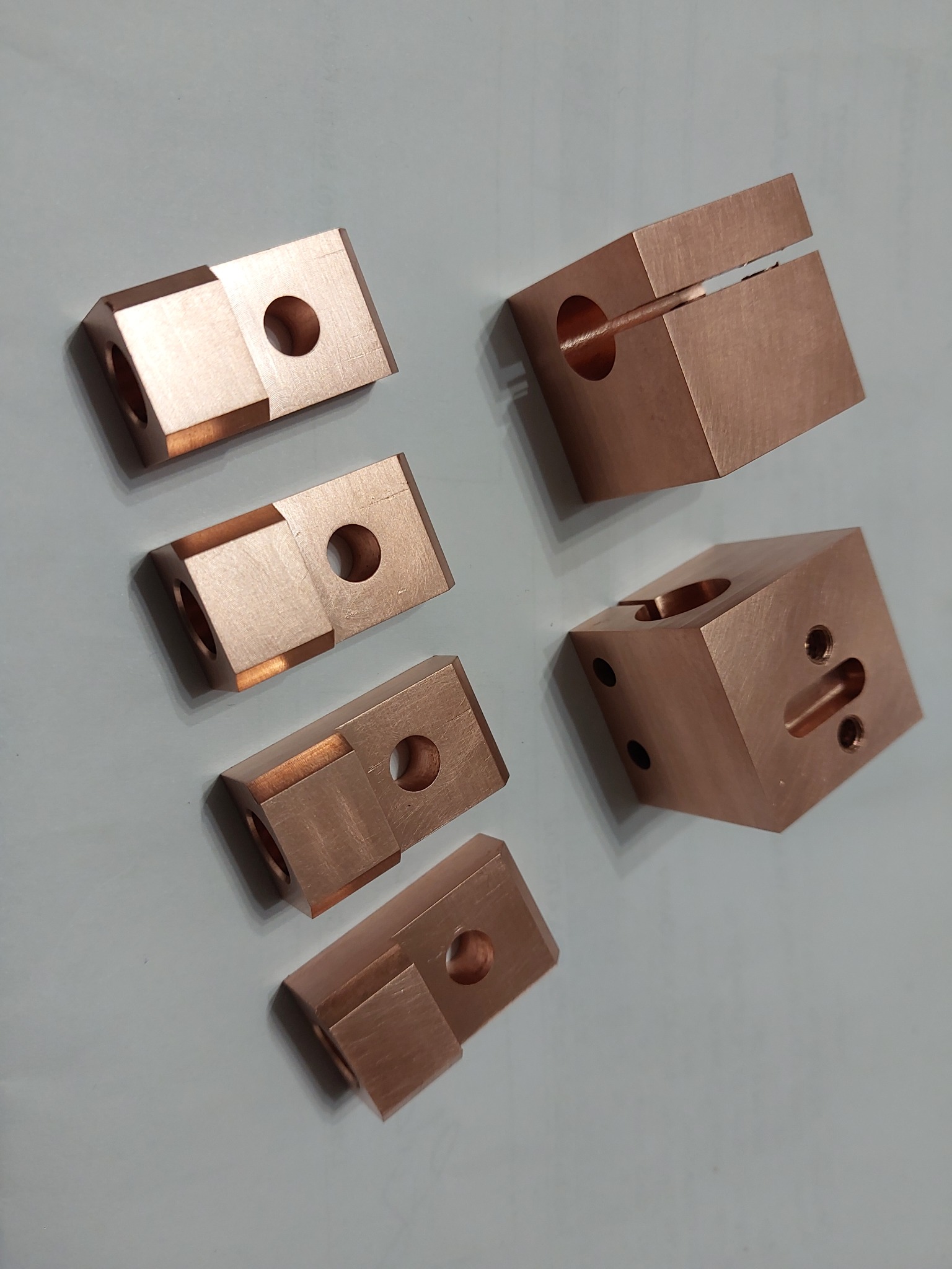 Miedziane, precyzyjnie obrobione elementy z otworami i rowkami, ułożone na szarym tle. Widoczne ślady obróbki skrawaniem i detale wykończenia.